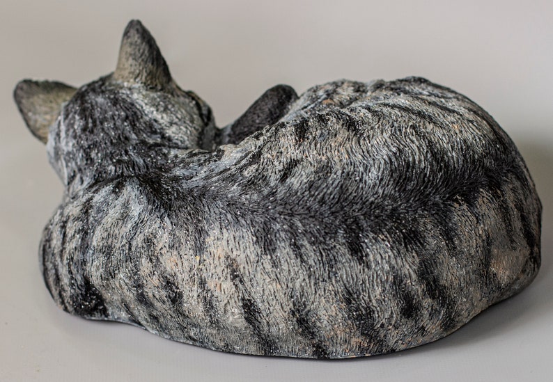 May include: A gray and white tabby cat sculpture curled up in a sleeping position. The sculpture is made of a textured material that resembles fur.