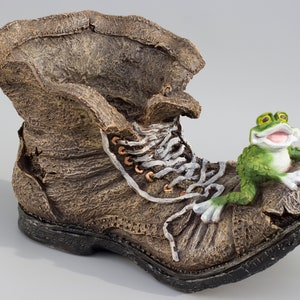 May include: A brown, worn-out boot with white laces and a green frog sitting on top. The frog is smiling and has a red ladybug on its back.