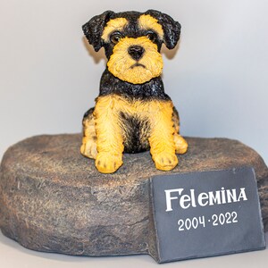 May include: A dog figurine with black and tan fur sits atop a stone-like base. The figurine has a detailed face and is posed in a seated position. A small plaque with the name "Felemina" and the years 2004-2022 is in front of the figurine.