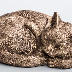 May include: A bronze-colored sculpture of a cat curled up sleeping. The cat's fur is detailed and realistic, and its eyes are closed. The sculpture is a great addition to any home decor.
