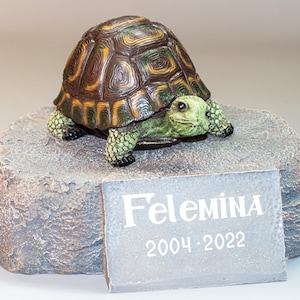 Puede incluir: Una pequeña figura de tortuga verde y marrón sobre una base de piedra gris con una placa que dice "Felemina 2004-2022".