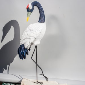 Könnte beinhalten: Eine weiße und blaue Keramik-Kranich-Statue mit rotem Schnabel und schwarzen Beinen. Der Kranich steht auf einem Sockel und hat den Kopf gesenkt.