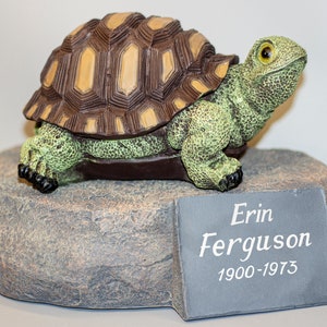 May include: A green and brown turtle figurine on a grey rock base with a black slate plaque that reads "Erin Ferguson 1900-1973".
