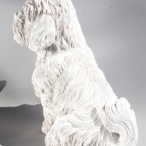 May include: White ceramic figurine of a sitting dog with fluffy fur. The dog is facing to the right and has its tail curled around its legs.