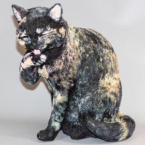 Puede incluir: Una figurita de cerámica de un gato con pelaje negro, marrón y blanco, sentado y lamiendo su pata.
