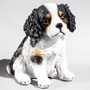 以下が含まれることがあります： 黒、白、茶色のキャバリア・キング・チャールズ・スパニエルの子犬の置物。置物は座って、頭を傾け、耳を立てています。