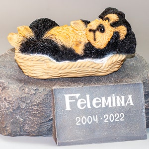 May include: A small black and brown dog figurine lying in a wicker basket on a gray stone base. A plaque on the base reads "Felemina 2004-2022".