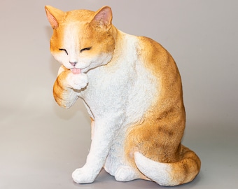May include: A white and orange cat figurine sitting on a gray surface. The cat is licking its paw and has its eyes closed.