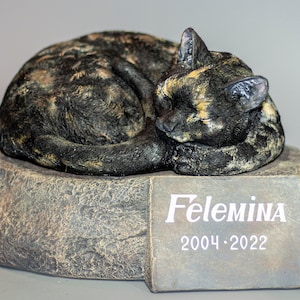 Puede incluir: Una figurita de gato negro, marrón y naranja durmiendo sobre una base de piedra gris con una placa que dice "Felemina 2004 - 2022".