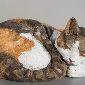 Może przedstawiać: Ceramiczna rzeźba śpiącego kota trójkolorowego zwiniętego w kłębek. Kot jest pomalowany w odcieniach brązu, pomarańczy, bieli i szarości.