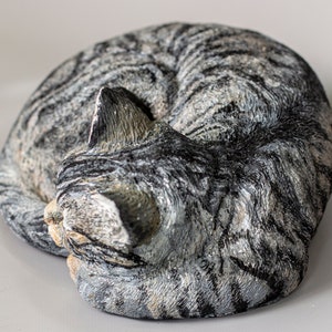 May include: A gray and white tabby cat sculpture curled up in a sleeping position. The sculpture is made of a textured material that resembles fur.