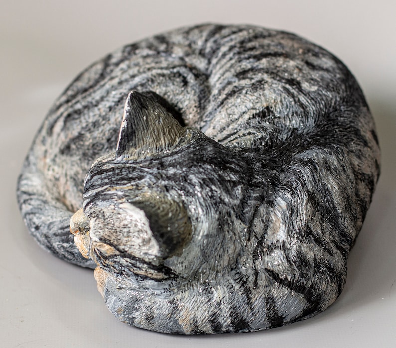 May include: A gray and white tabby cat sculpture curled up in a sleeping position. The sculpture is made of a textured material that resembles fur.