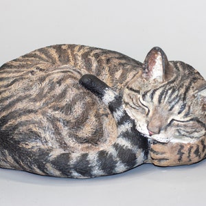 Puede incluir: Una escultura de cerámica de un gato atigrado enroscado y durmiendo. El gato está pintado en tonos marrones, negros y blancos, con detalles realistas.
