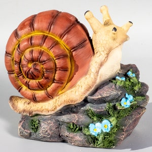 May include: A brown and yellow snail figurine with a detailed shell, crawling on a rock with blue flowers. The snail is made of resin and is a garden decoration.