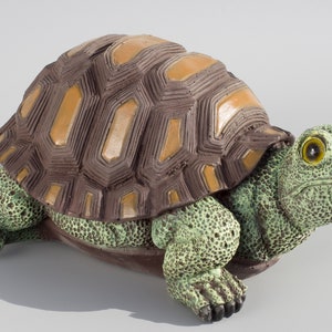 Puede incluir: Una figurita de tortuga de cerámica con una concha marrón y naranja y un cuerpo verde. La tortuga tiene una concha texturizada detallada y una cara realista con ojos grandes.