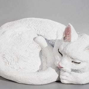Peut inclure: Une sculpture en céramique blanche d'un chat enroulé en train de dormir. Le chat a des oreilles roses et une texture de fourrure réaliste et détaillée.