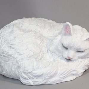 May include: White ceramic sculpture of a cat curled up sleeping. The cat has a fluffy, detailed coat and closed eyes.