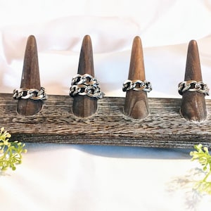 May include: Six silver chain rings displayed on individual wooden ring holders. The rings are on a rustic wooden base with a white background. Greenery is placed on the base in the foreground.