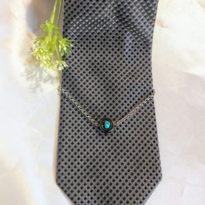 May include: A silver tie clip with a turquoise stone set in a black circle. The tie clip is on a gray patterned tie.