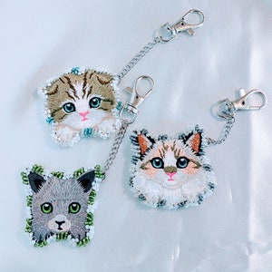 May include: Three embroidered cat keychains with silver chains. The keychains feature different cat breeds with white, brown, and gray fur.