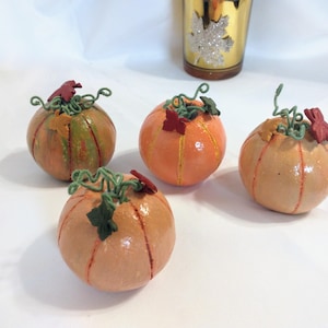 May include: Four small, decorative pumpkins in shades of orange and brown, with green vines and red leaf accents. The pumpkins are arranged on a white surface, with a gold-colored glass in the background. The pumpkins are a fall-themed decorative item.