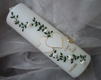 Puede incluir: Vela de boda blanca con dos corazones dorados y una vid de rosas blancas y hojas verdes. La vela está inscrita con los nombres "Cintia & Hilgar" y la fecha "28.08.2021".
