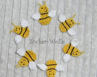 May include: Six crocheted bumblebees with black and yellow stripes and white wings are arranged in a circle on a white fuzzy surface. The text "Zucker-Wolle" is in the center of the circle.