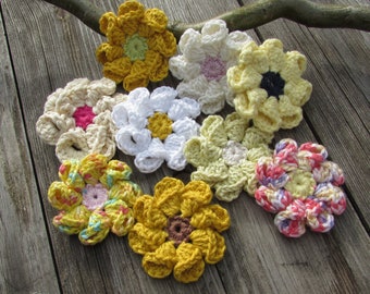 Puede incluir: Una colección de ocho flores de ganchillo en varios colores, incluyendo amarillo, blanco, rosa y morado. Las flores están dispuestas sobre una superficie de madera. El texto "Zucker-Wolle" es visible en la parte inferior de la imagen.