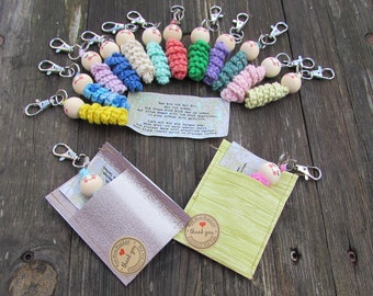 Puede incluir: Una colección de pequeños llaveros con cabezas de cuentas de madera con caras pintadas y cuerpos de ganchillo de colores. Dos llaveros están metidos en pequeñas bolsas de tela con pegatinas de 'thank you'. Se incluye una nota con texto en alemán.