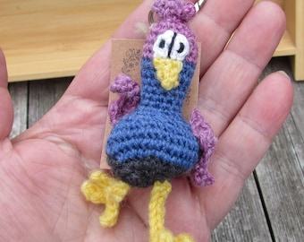 Puede incluir: Un llavero de ganchillo con forma de criatura parecida a un pájaro azul y morado, con pico y patas amarillas, ojos blancos con pupilas negras y pequeñas alas moradas. La figura está unida a una anilla de metal plateado sobre una etiqueta.