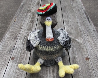 Puede incluir: Una figura de pavo tejida a mano con cuerpo gris oscuro, patas amarillas y cuello blanco. El pavo lleva una boina colorida con círculos concéntricos rojos, amarillos, verdes y negros. Tiene ojos blancos con pupilas negras y un pico amarillo.