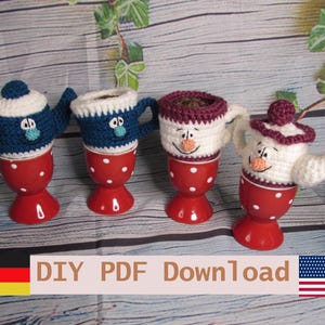 May include: Four red egg cups with white polka dots, each topped with a crocheted cozy. Two cozies are shaped like teapots in blue and white, while the other two are mug-shaped in blue and white or maroon and white, featuring embroidered faces.