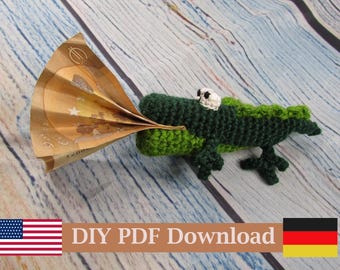 Puede incluir: Un juguete de cocodrilo tejido a ganchillo en verde oscuro y verde claro con ojos blancos. El cocodrilo tiene la boca abierta y sostiene un billete de cinco euros marrón doblado. El texto DIY PDF Download es visible abajo.