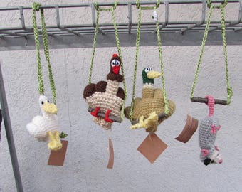 Puede incluir: Cuatro adornos de animales tejidos a ganchillo colgados de una rejilla metálica con cordones trenzados verdes. Las figuras incluyen un pájaro blanco, un pavo marrón, un pato azulón y una zarigüeya gris colgada por la cola.