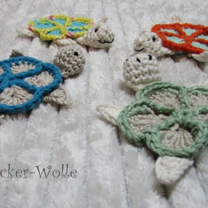 Könnte beinhalten: Fünf gehäkelte Schildkröten-Anhänger in verschiedenen Farben, darunter Blau, Grün, Orange und Gelb. Die Schildkröten liegen auf einer weißen, flauschigen Oberfläche. Der Text "Zucker-Wolle" ist in der linken unteren Ecke des Bildes sichtbar.