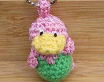 Puede incluir: Un llavero de ganchillo que representa una pequeña criatura rellena con cabeza rosa, pico amarillo y cuerpo verde. La figura tiene dos pequeños ojos negros y está unida a una anilla de metal plateado en la parte superior para colgarla.