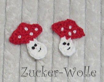Puede incluir: Dos apliques de setas de ganchillo hechos a mano. Cada seta tiene un sombrero rojo con puntos blancos y un tallo blanco con una cara sonriente. El texto "Zucker-Wolle" está en la parte inferior. Ideal para proyectos de manualidades.