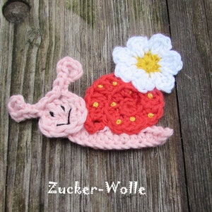 Könnte beinhalten: Eine gehäkelte Schnecke mit Erdbeerschale und Blume auf dem Rücken. Die Schnecke ist rosa, die Schale rot mit gelben Punkten und die Blume weiß mit gelbem Zentrum. Der Text "Zucker-Wolle" befindet sich unten.