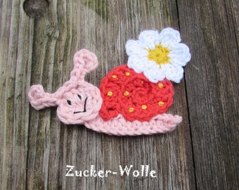 Puede incluir: Un caracol de ganchillo con una concha de fresa y una flor en la espalda. El caracol es rosa, la concha es roja con puntos amarillos y la flor es blanca con un centro amarillo. El texto "Zucker-Wolle" está en la parte inferior.