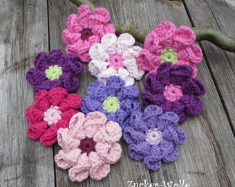 Puede incluir: Siete flores de ganchillo en varios tonos de rosa, morado y verde. Las flores están dispuestas en círculo sobre una superficie de madera. El texto "Zucker-Wolle" es visible en la esquina inferior derecha de la imagen.