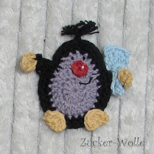 Puede incluir: Un gnomo de crochet morado y negro con una nariz roja y un lazo de crochet azul y blanco. El gnomo sostiene una flor de crochet azul y blanco. El texto "Zucker-Wolle" es visible en la esquina inferior derecha de la imagen.