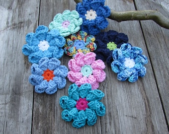 Puede incluir: Una colección de ocho flores de ganchillo en varios colores, incluyendo azul, rosa, verde y amarillo. Las flores están dispuestas sobre una superficie de madera.