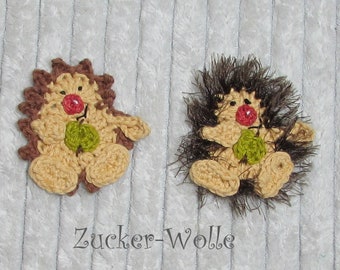 Puede incluir: Dos erizos de crochet marrón y beige con narices rojas y hojas verdes. Los erizos están sentados sobre una superficie blanca y peluda. El texto "Zucker-Wolle" está escrito debajo de los erizos.