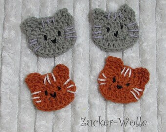 May include: Four crocheted cat faces, two grey and two orange, with black eyes and white whiskers. The grey cats have purple yarn for their whiskers, and the orange cats have white yarn for their whiskers.