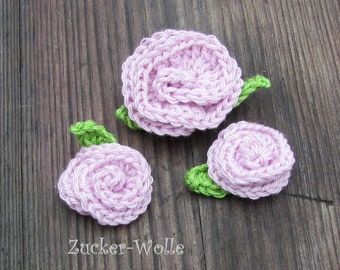 Puede incluir: Tres rosas de crochet de color rosa con hojas verdes. Las rosas son de diferentes tamaños y están dispuestas sobre una superficie de madera.