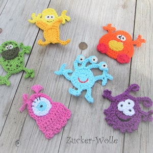 Könnte beinhalten: Sechs gehäkelte Monsterfiguren in verschiedenen Farben, darunter grün, gelb, orange, blau, rosa und lila. Jedes Monster hat ein einzigartiges Design und große Kulleraugen.