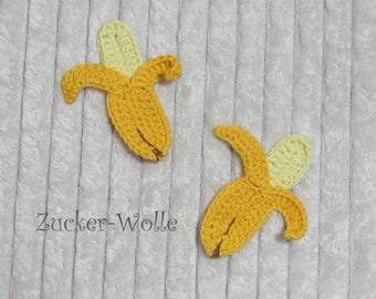 Puede incluir: Dos formas de plátano de crochet, una amarilla y otra amarillo claro, sobre un fondo blanco peludo. El texto "Zucker-Wolle" es visible debajo de los plátanos.