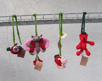 Puede incluir: Cuatro adornos de ganchillo colgados de una rejilla metálica. Los artículos incluyen una fresa roja, una mariposa marrón y rosa, un caracol rosa claro con una concha de fresa y una flor blanca, y un oso de peluche rojo, cada uno con etiqueta.