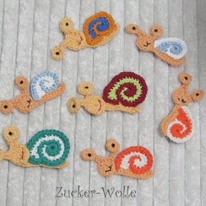 Könnte beinhalten: Eine Sammlung handgefertigter Häkelapplikationen in Form von Schnecken. Jede Schnecke hat einen beigefarbenen Körper und ein Schneckenhaus in Blau, Orange, Grün und Rot. Die Applikationen sind mit detaillierten Spiralmustern und kleinen, runden Augen versehen.