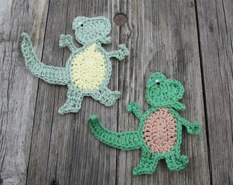 Puede incluir: Dos apliques de dinosaurios hechos a mano con ganchillo. Un dinosaurio es verde claro con vientre amarillo, el otro es verde con vientre marrón. Ambos tienen ojos pequeños y redondos y están posados con los brazos y la cola extendidos.